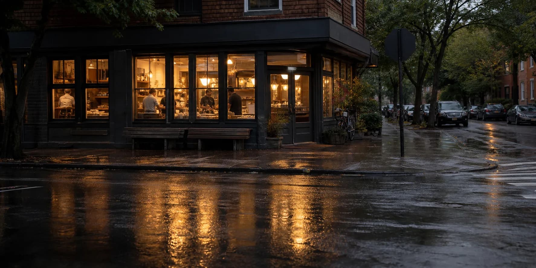 An independent corner shop at dusk seen from across a quiet street, its windows the only lit thing on the block
